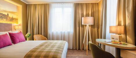 Ein Hotelzimmer mit Bett, Schreibtisch, Stuhl, Lampe und einem fensterbepflanzten Fenster.