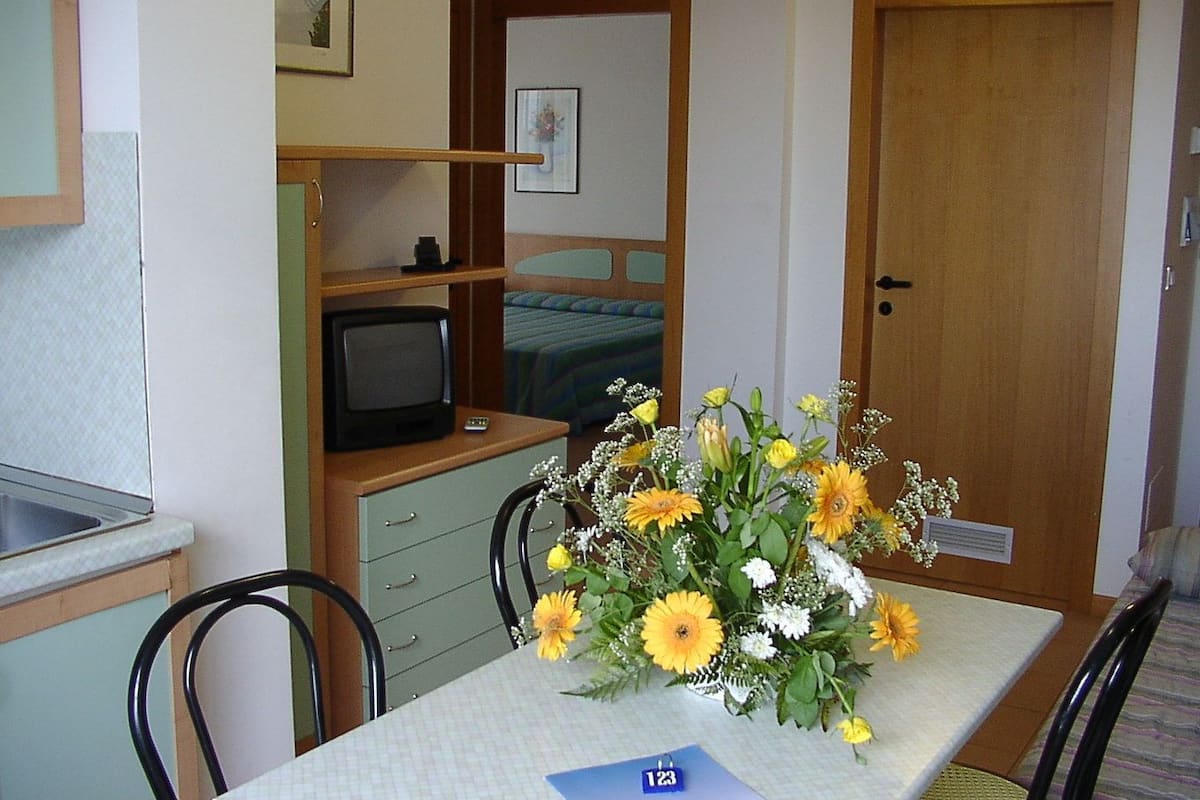 Ein kleines Zimmer mit einem Bett, einem Fernseher auf einem Regal, einem Tisch mit Blumenvase und einer Tür.
