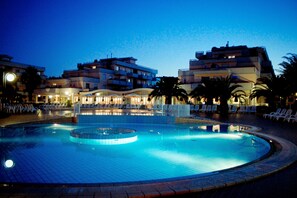 Une piscine entourée de bâtiments dotés de balcons et d’éclairages, avec des chaises longues et des parasols à proximité.