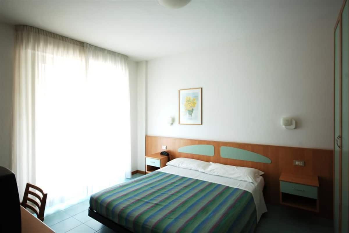 Ein Hotelzimmer mit Bett, Nachttisch, Stuhl, Schreibtisch und einem Fenster mit Vorhängen.