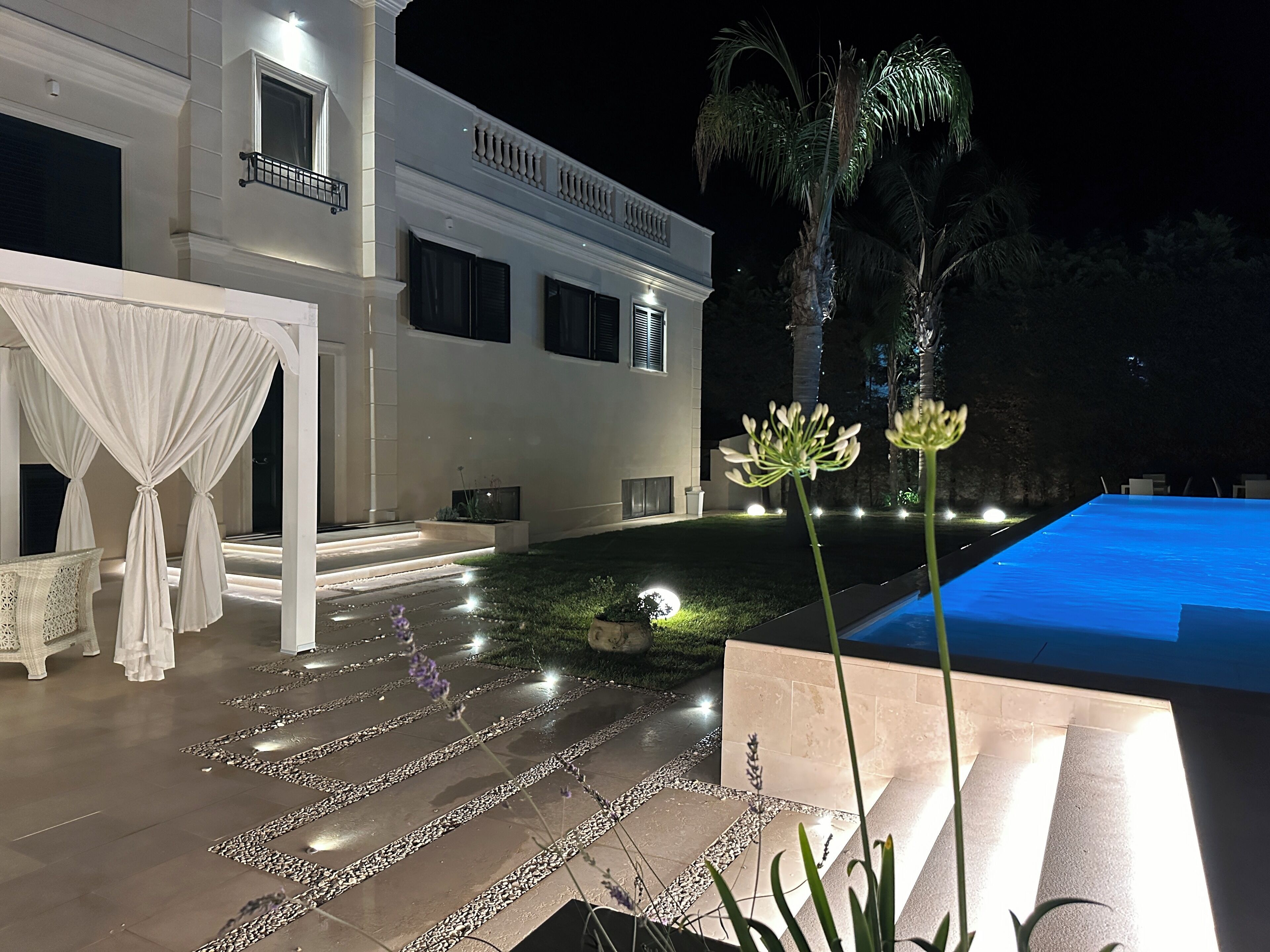 A modern outdoor pool area with a well-lit patio, a white building with large windows, and a neatly landscaped garden with a pathway leading to the pool.