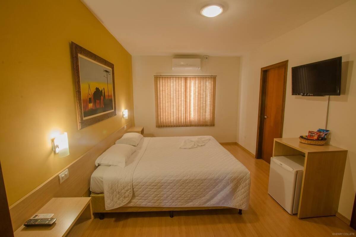 Une chambre d’hôtel comprenant un lit, une télévision, un petit bureau, un climatiseur fixé au mur et une fenêtre avec des rideaux.