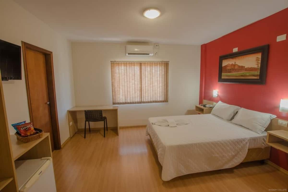Une chambre à coucher comprenant un lit, un bureau, une chaise, une fenêtre avec des stores, une table de chevet et un climatiseur fixé au mur.