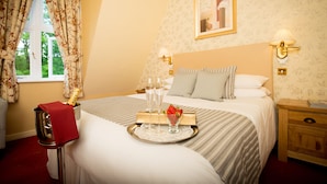 Una habitación de hotel con cama, mesita de noche con lámpara, una bandeja con copas de champán y fresas, y una ventana con cortinas.