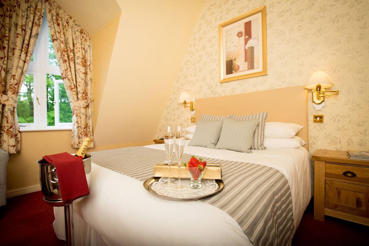 Een hotelkamer met een bed, een nachtkastje met een lamp, een dienblad met champagneglazen en aardbeien, en een raam met gordijnen.