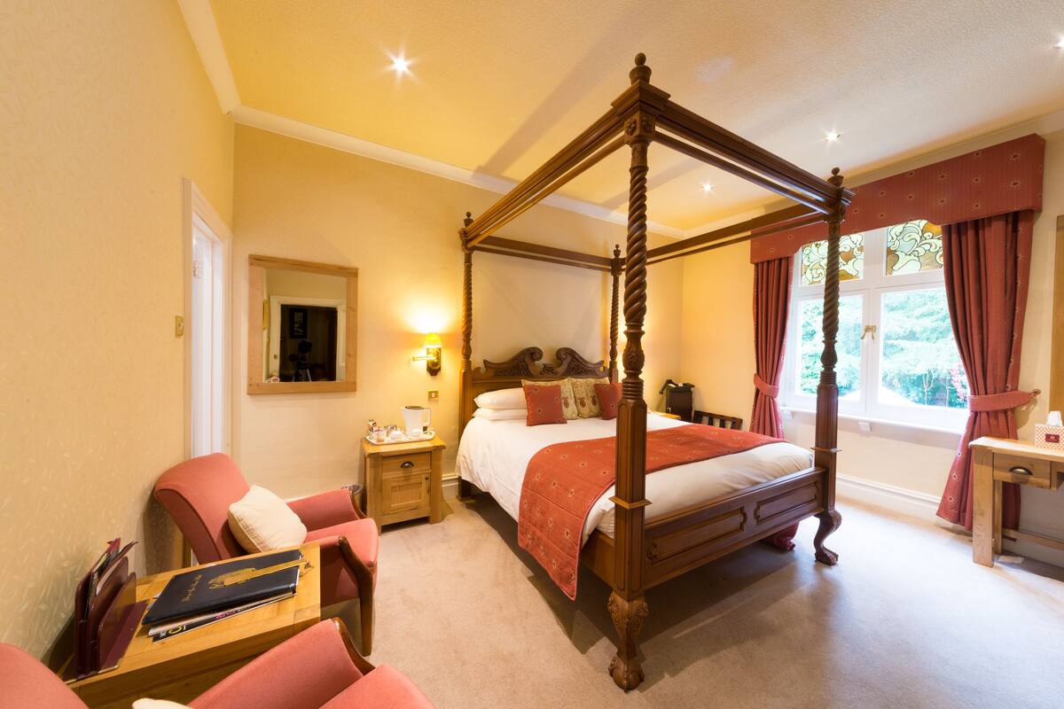 Een bed met hemelbed en houten frame, een nachtkastje met lamp en een stoel met kussen.