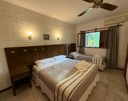 Habitación de hotel con pared de ladrillo, dos camas, ventilador de techo, ventana con cortinas y un equipo de aire acondicionado.