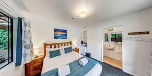 A bedroom with a bed, a nightstand, a painting, a bathroom with a sink and mirror, and a view of greenery outside. - Lochmara Lodge (Lochmara Bay)