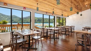A restaurant with wooden tables and chairs, large windows with a view of a lake and mountains, and hanging lanterns. - Lochmara Lodge (Lochmara Bay)