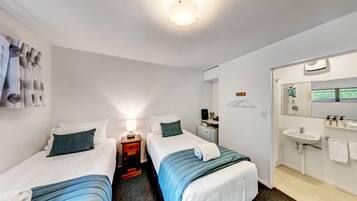 Habitación de hotel con dos camas individuales, una mesita de noche con lámpara, un escritorio pequeño con silla, un baño con lavamanos y espejo, y una ventana con cortinas.