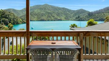 A wooden deck with a table and two chairs, overlooking a turquoise lake and green hills.