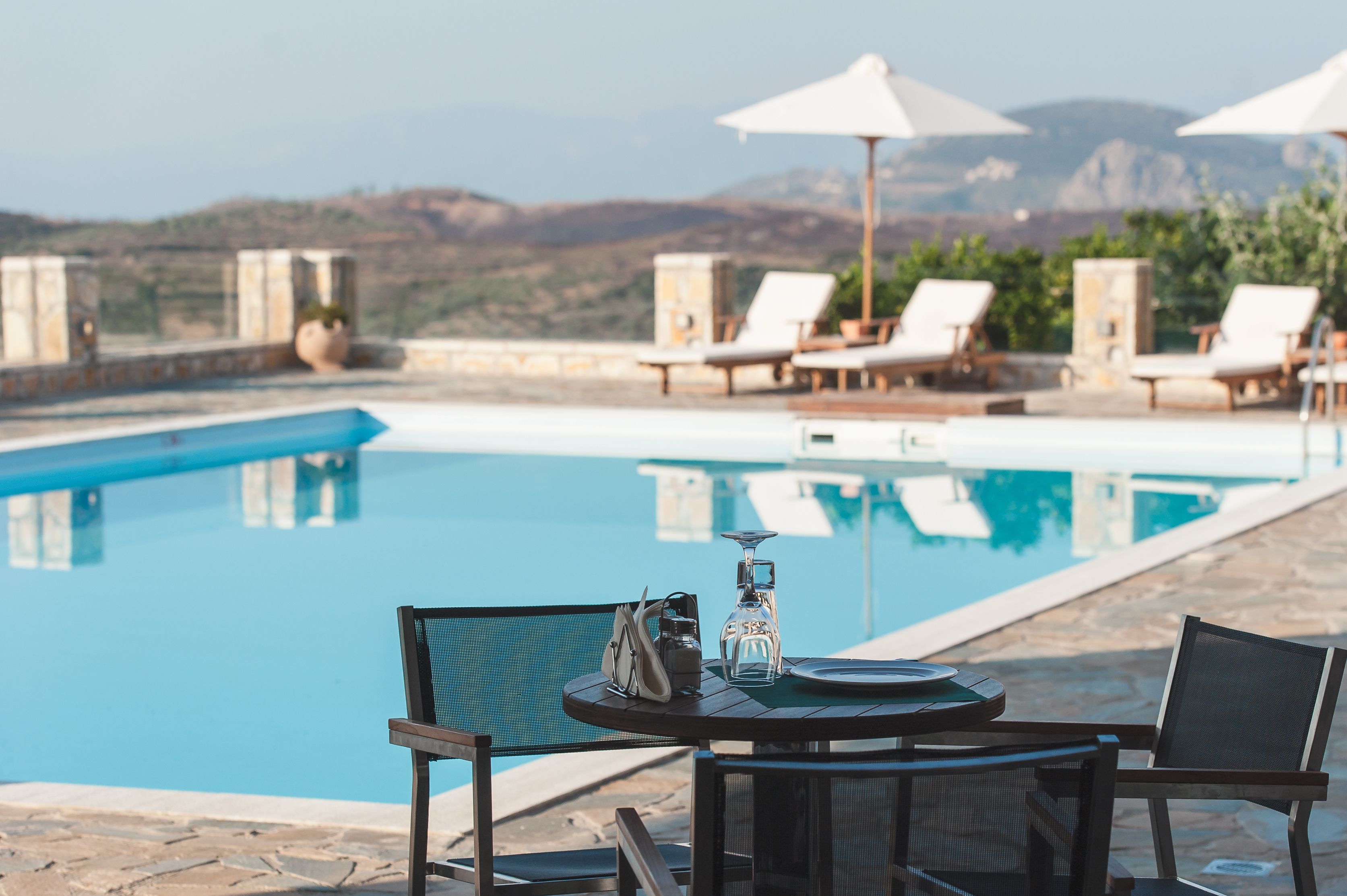 Zona de piscina con sillones de descanso y sombrillas, una mesa con una botella y vasos, y vistas a montañas al fondo.