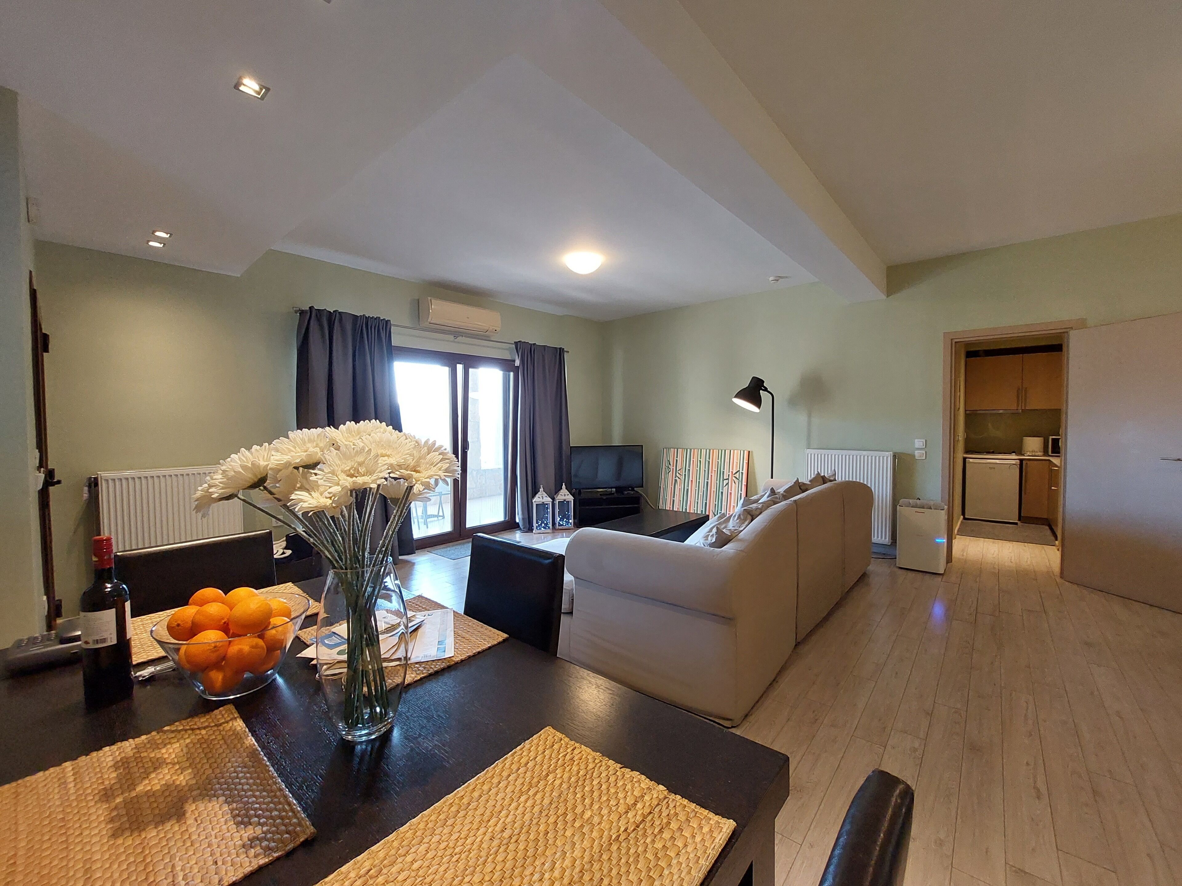 Ein modernes Wohnzimmer mit einer Couch, einem Esstisch mit Blumenvase und einem durchgehenden Gang sichtbaren Küchenbereich.