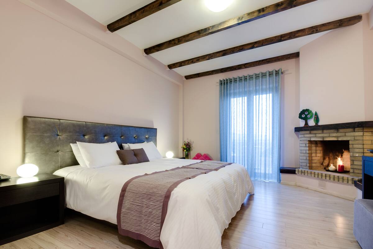 A bedroom with a large bed, wooden beams on the ceiling, a fireplace, and a nightstand with a lamp.