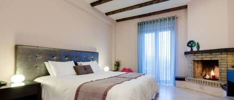 A bedroom with a large bed, wooden beams on the ceiling, a fireplace, and a nightstand with a lamp.