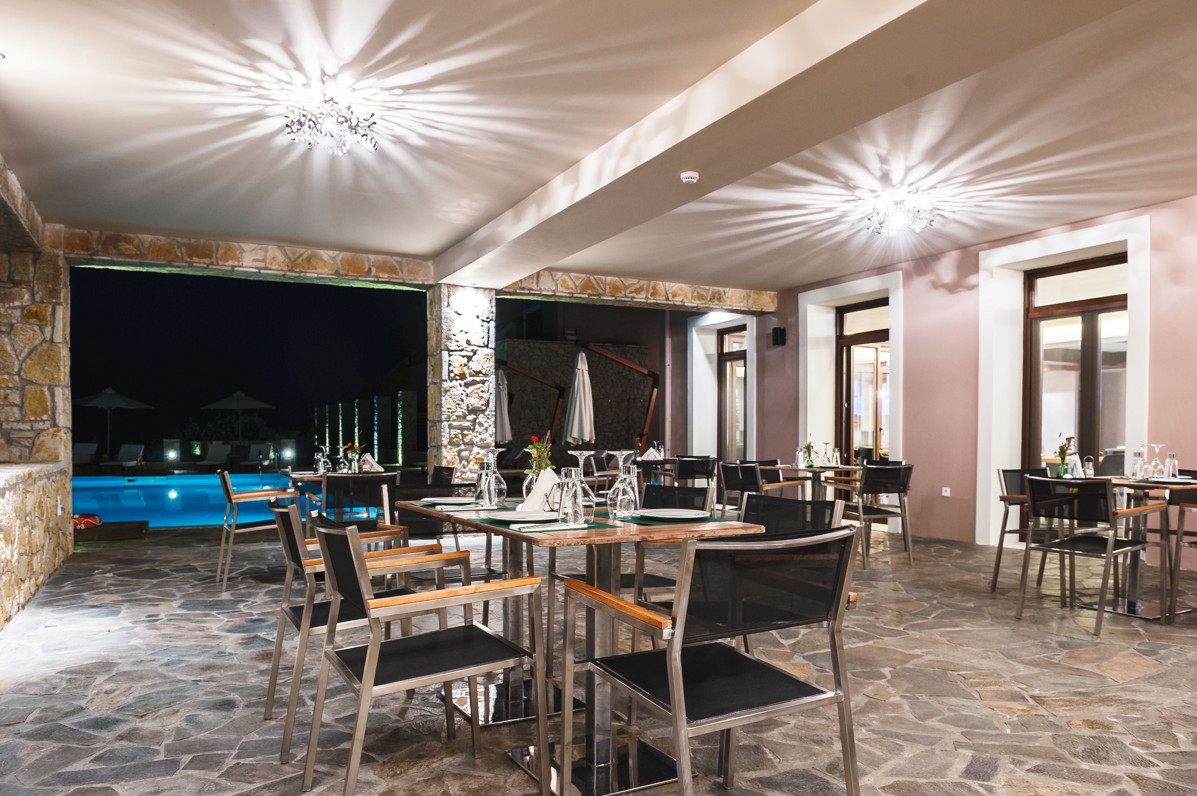 An outdoor dining area with stone flooring, tables set for dining, and a pool in the background.
