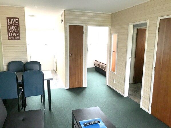 Une pièce comprenant une table à manger et des chaises, un canapé et une petite table avec une télécommande. Deux portes donnent accès à d’autres pièces.