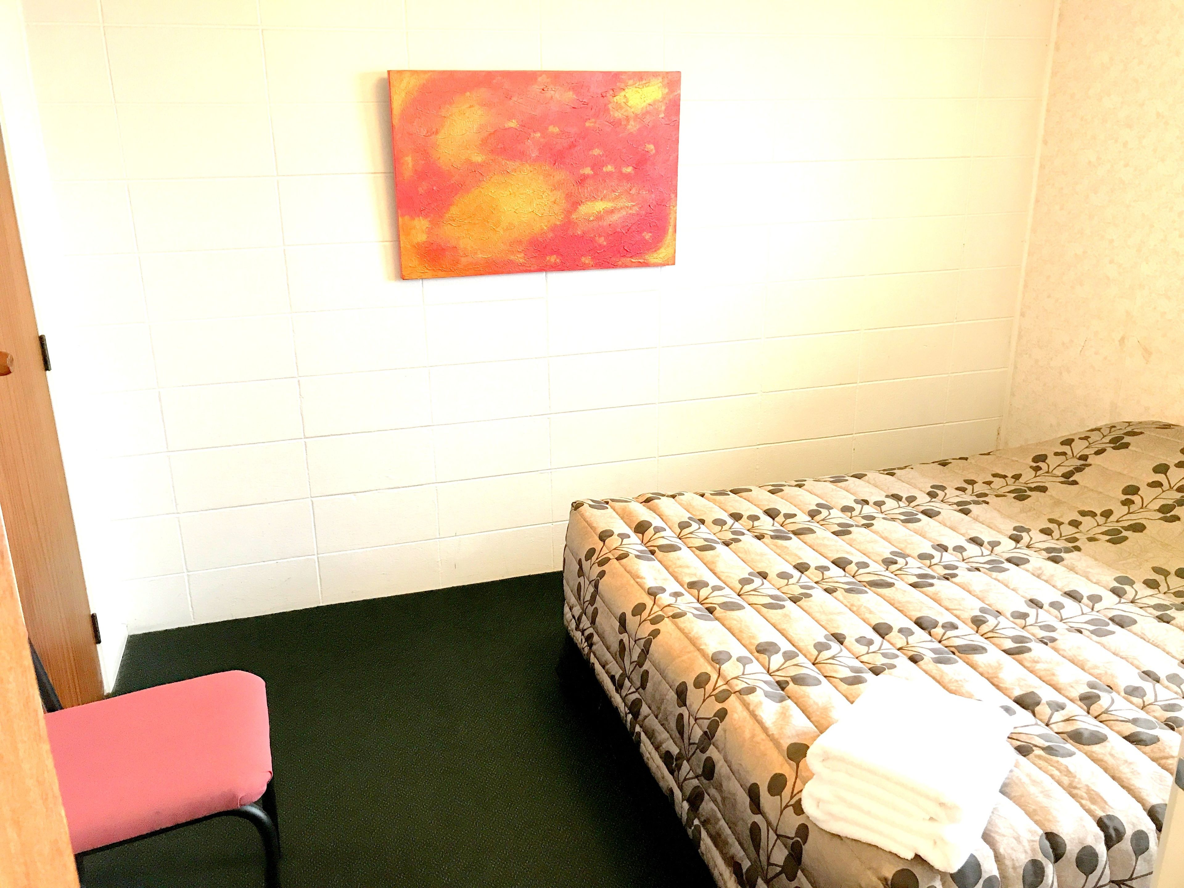 A small, simple room with a bed, a chair, and a wall-mounted painting.