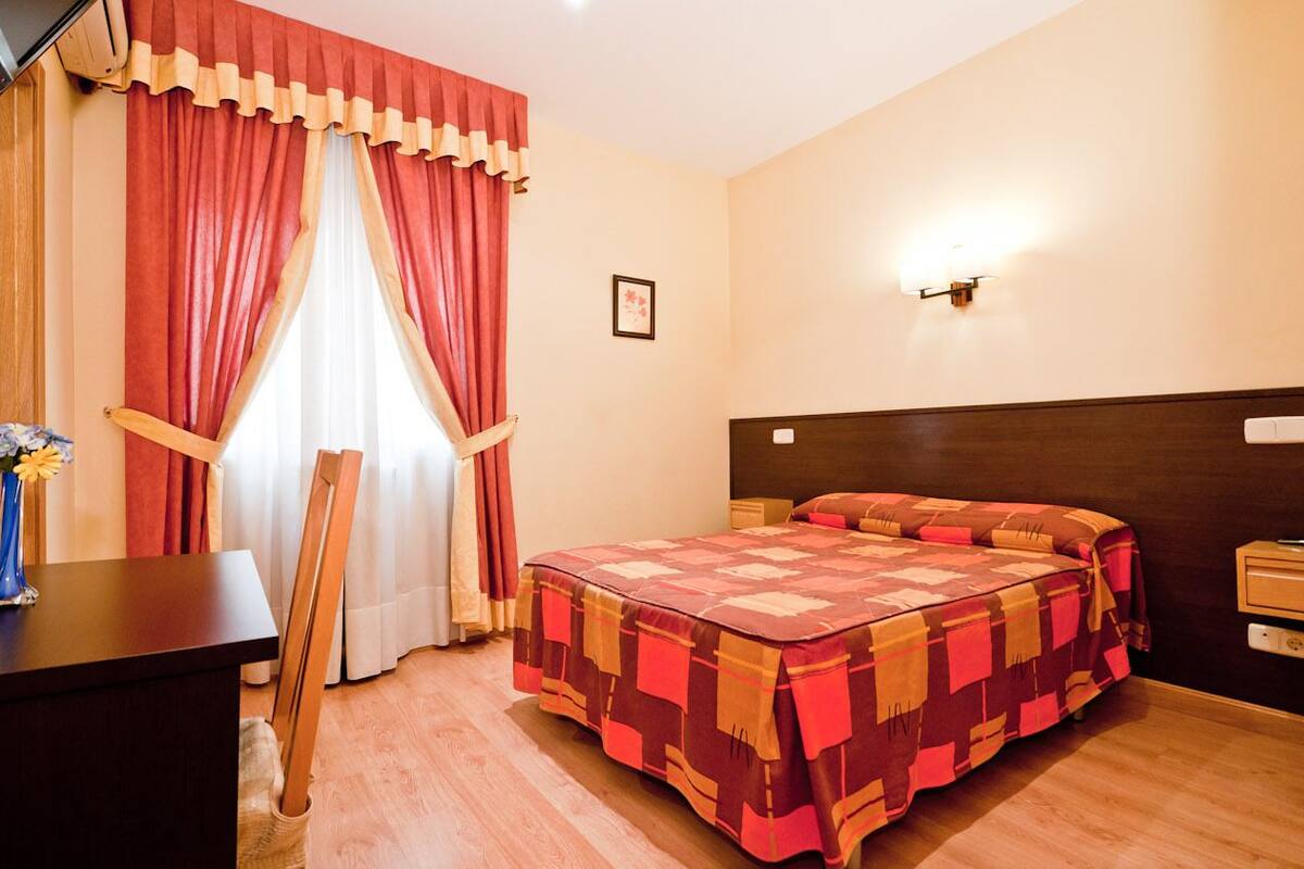 Habitación de hotel con cama, escritorio, silla, mesita de noche y lámpara de pared.