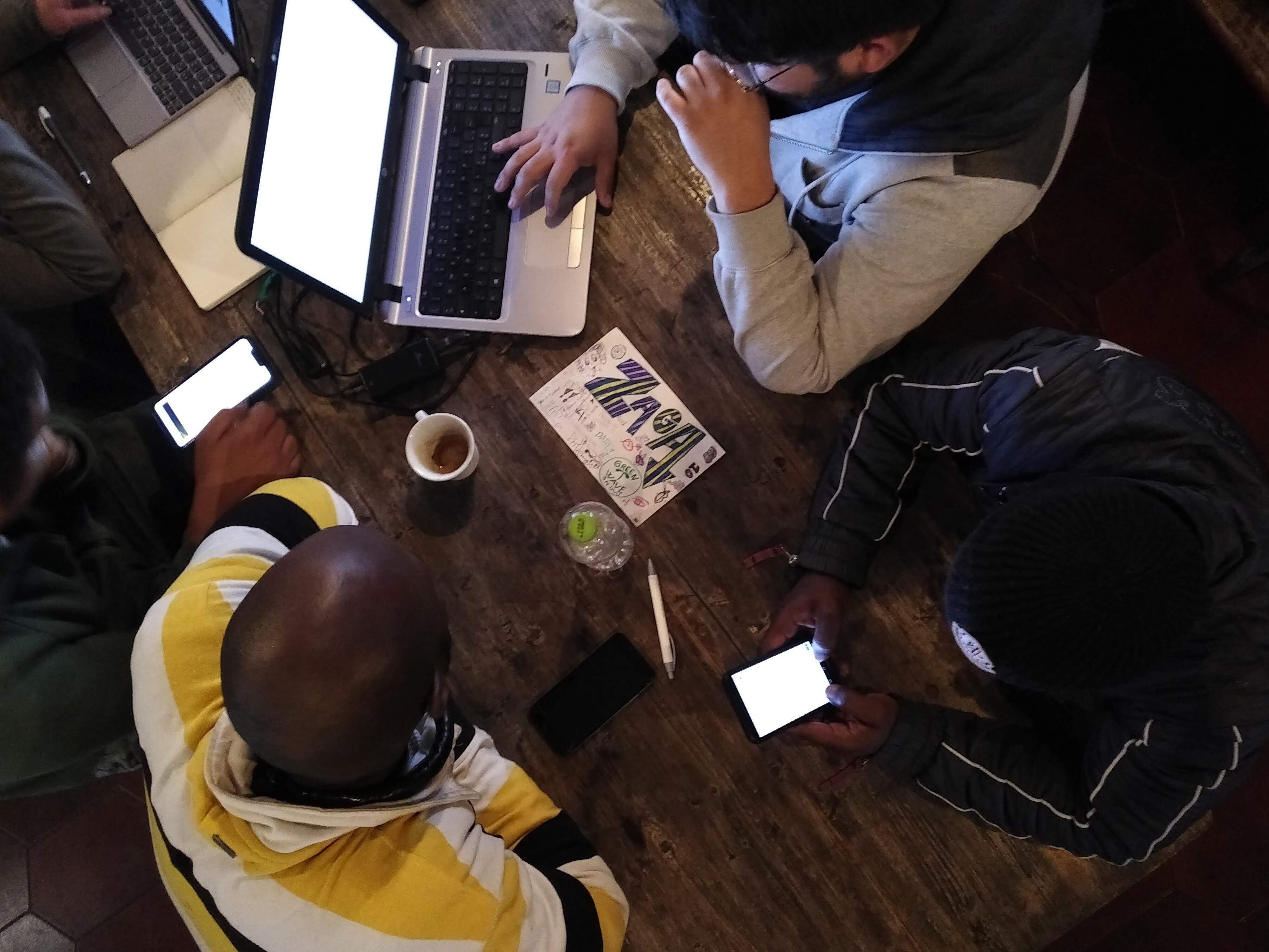 A group of people sitting around a wooden table with laptops and smartphones, a notebook, and a mug.