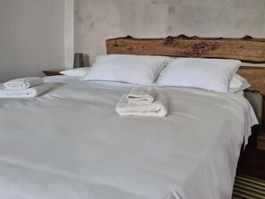 Una cama bien hecha con sábanas y toallas blancas, un cabecero de madera y una pared lisa.