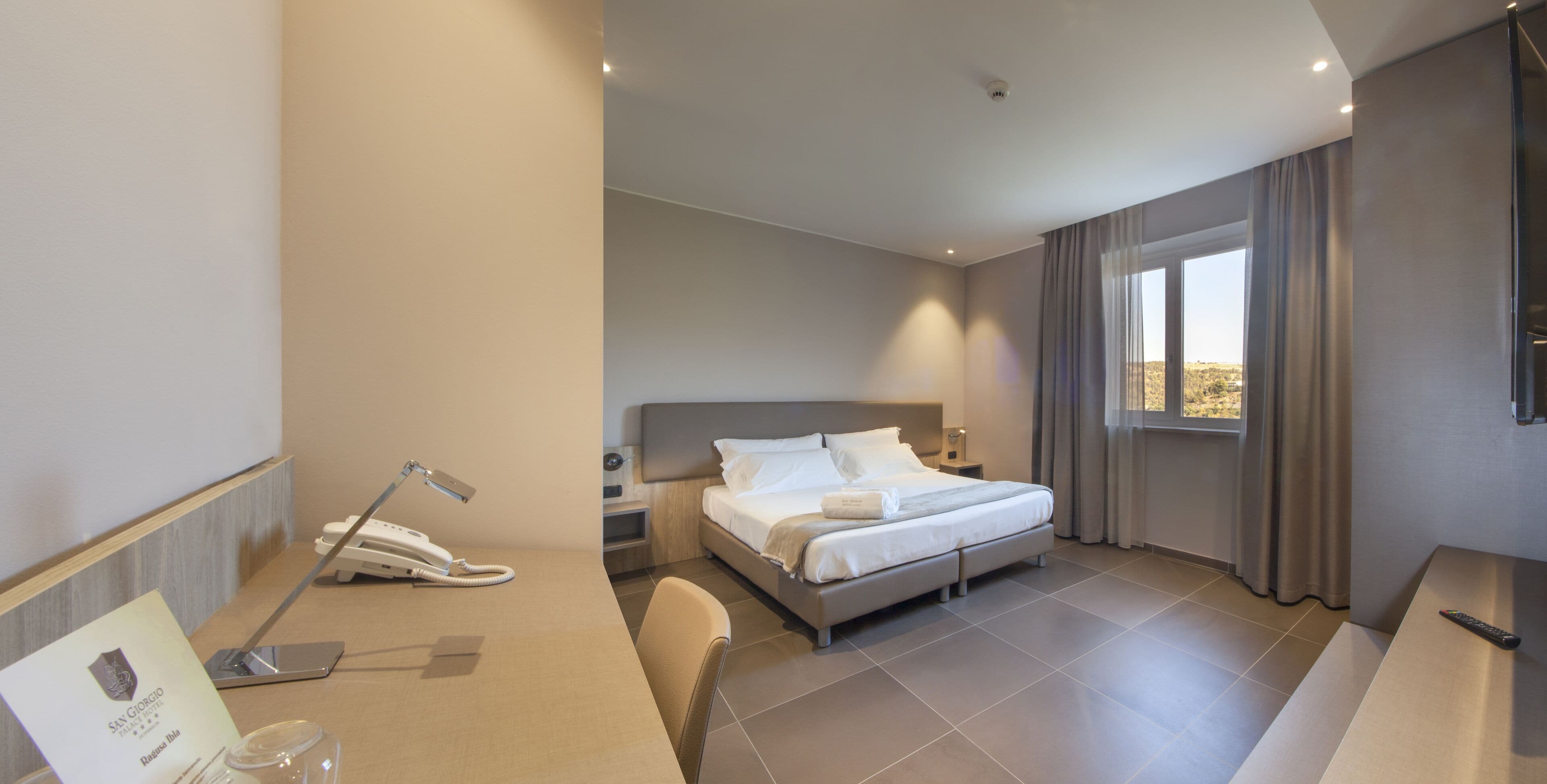 Een moderne hotelkamer met een bed, nachtkastje, lamp en een bureau met telefoon.