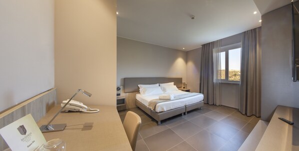 A modern hotel room with a bed, bedside table, lamp, and a desk with a telephone.