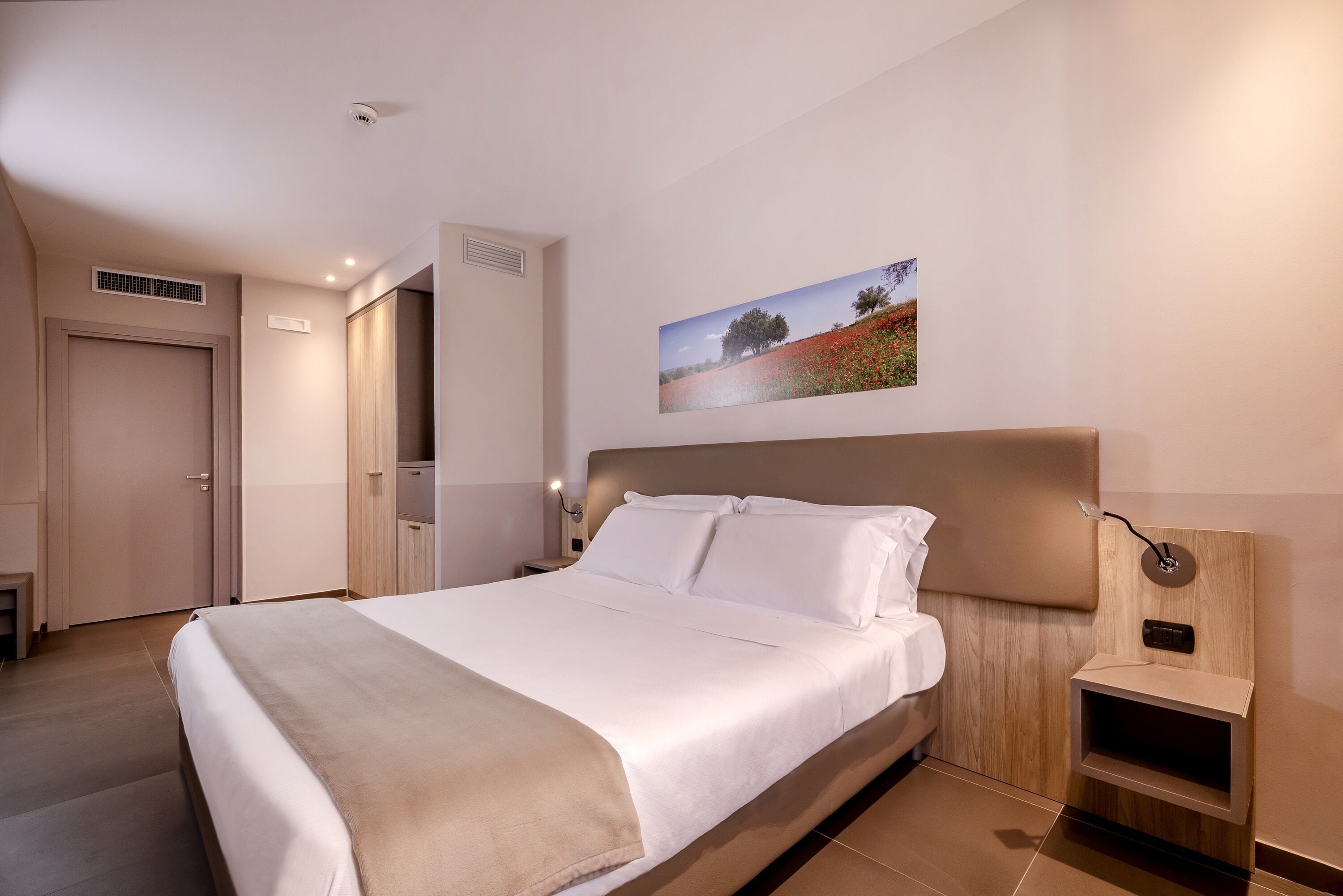 Una habitación de hotel moderna con una cama grande, una mesita de noche, una lámpara de pared y un cuadro de un paisaje.