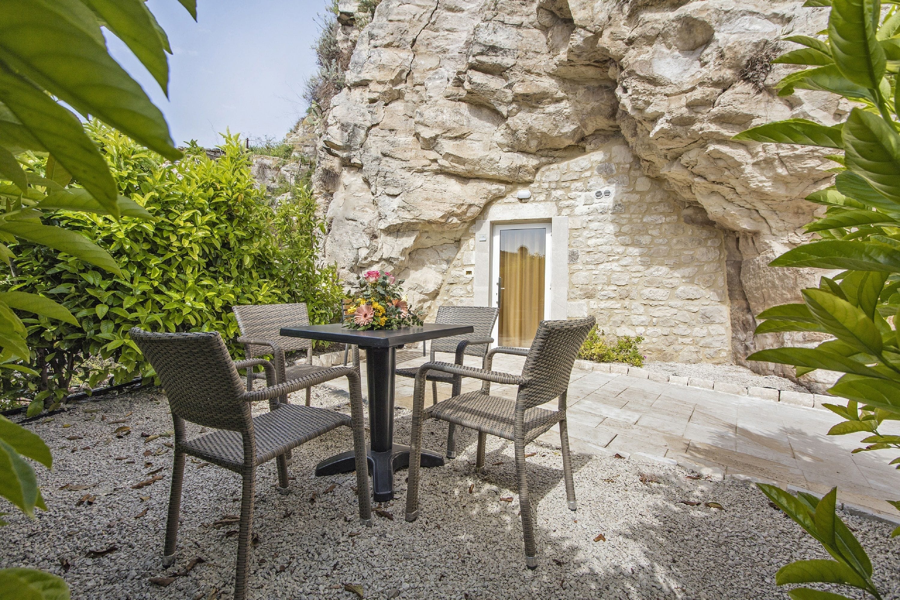 An outdoor seating area with wicker chairs and a table, set against a stone wall with a door.