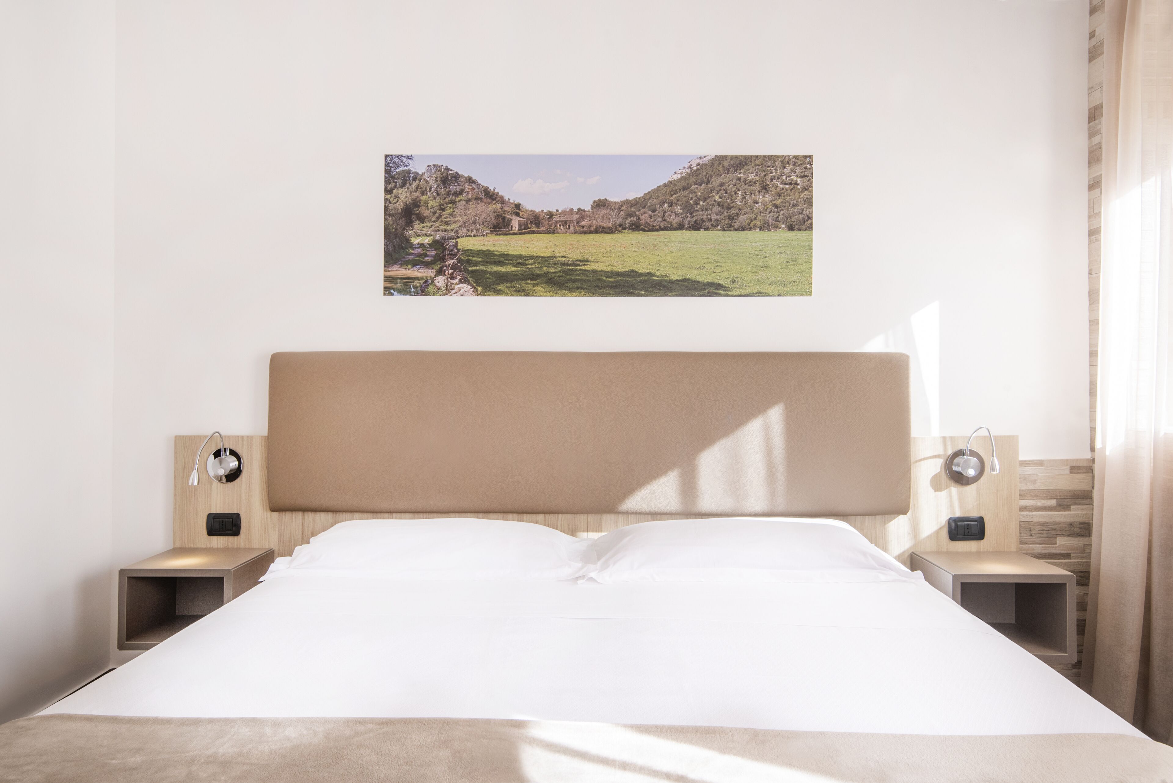 A neatly made bed with a headboard, two bedside tables with lamps, and a framed picture of a landscape on the wall.