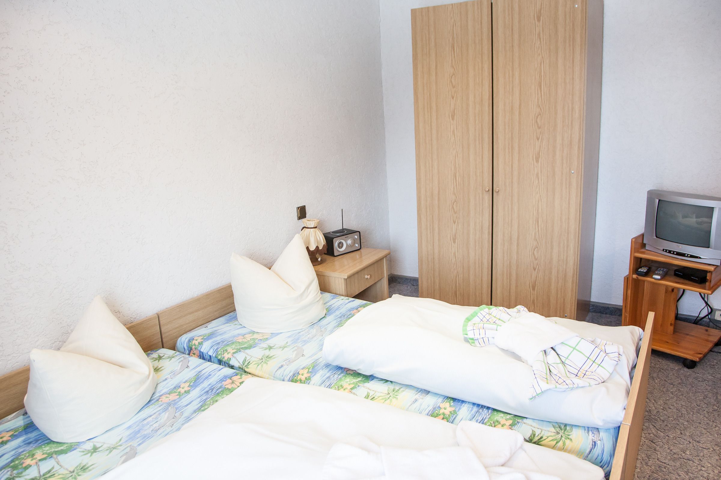 Una camera d'albergo con due letti, un'armadio in legno, un comodino con una radio e un televisore su un supporto.