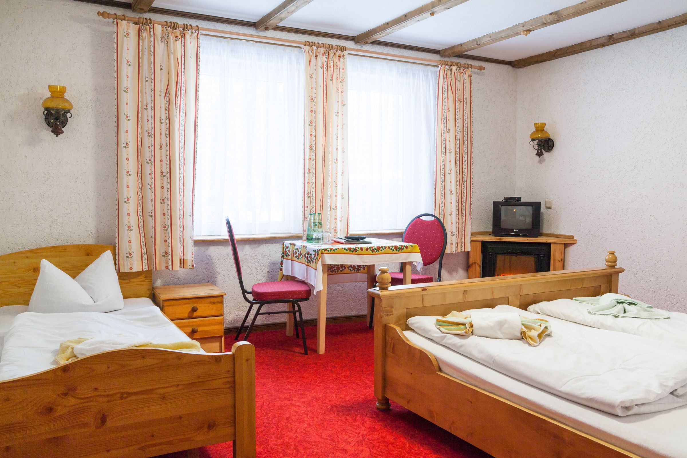 Una habitación con dos camas de madera, una mesita con una silla, televisión y una ventana con cortinas.