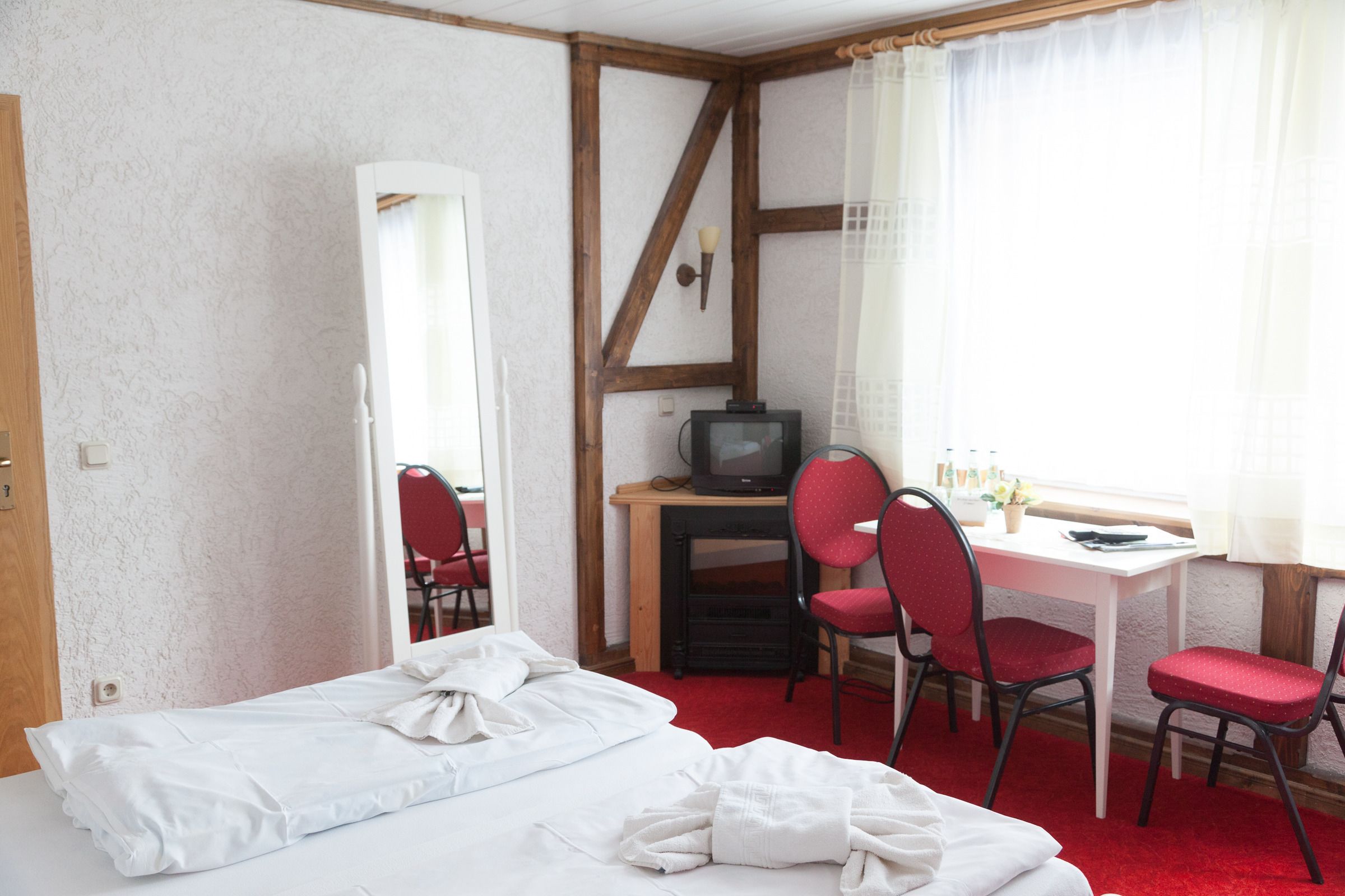 Ein Zimmer mit einem Bett, einem Schreibtisch mit Computer, zwei roten Stühlen, einem Spiegel und einem fenster mit Vorhängen.