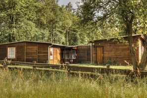 A rustic cabin with a porch, surrounded by tall trees and overgrown grass. - Pension Waldoase Hirschbüchenkopf (Harzgerode)