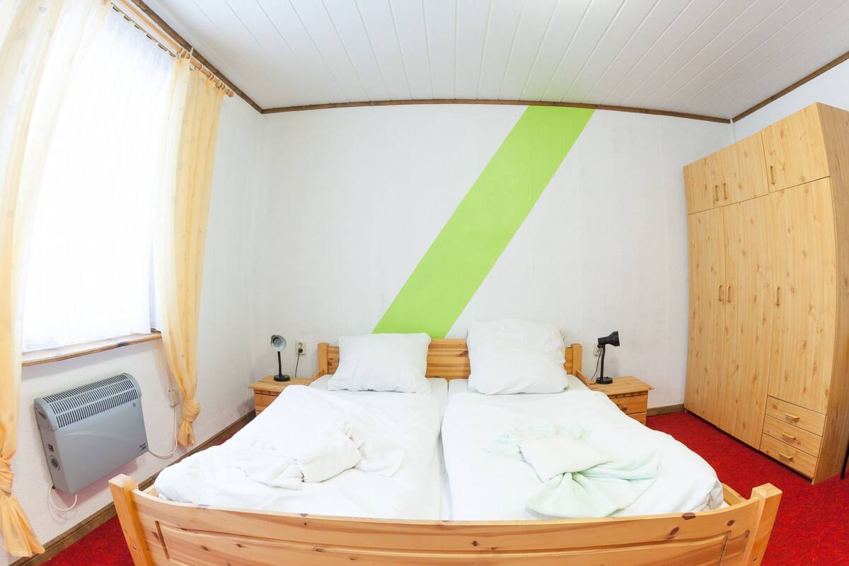 A bedroom with a wooden bed, two bedside tables, a green diagonal stripe on the wall, a wooden wardrobe, and a radiator.