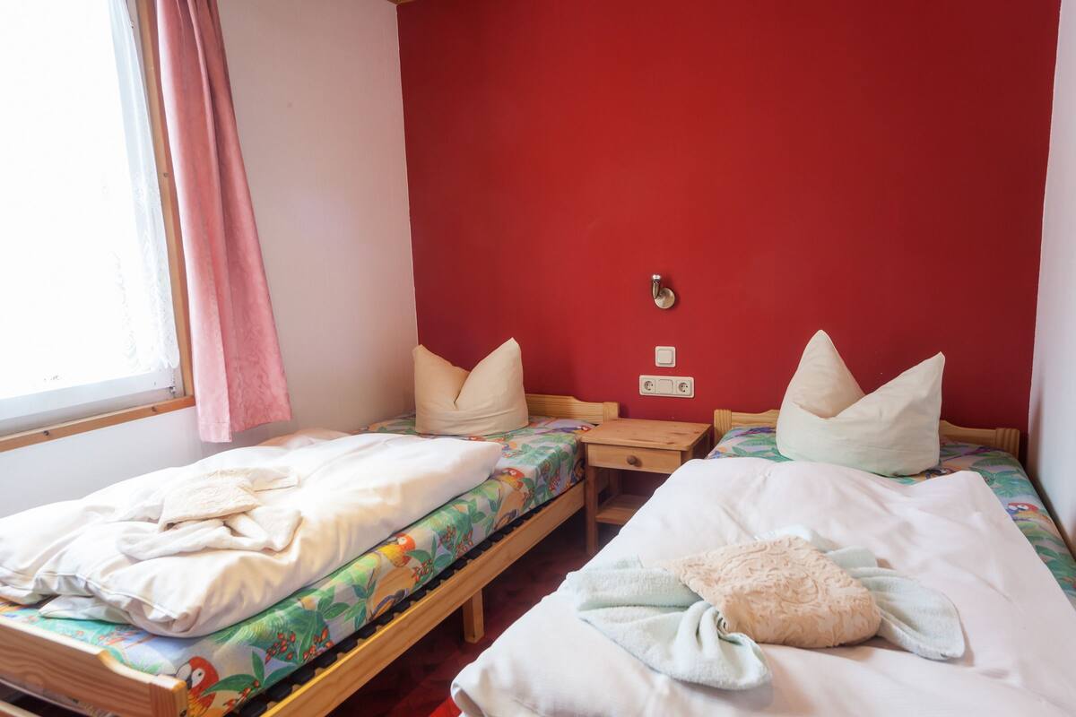 A room with two single beds, each with a pillow and a blanket. There is a small wooden nightstand between the beds. The walls are painted red, and there is a window with a curtain on the left side.