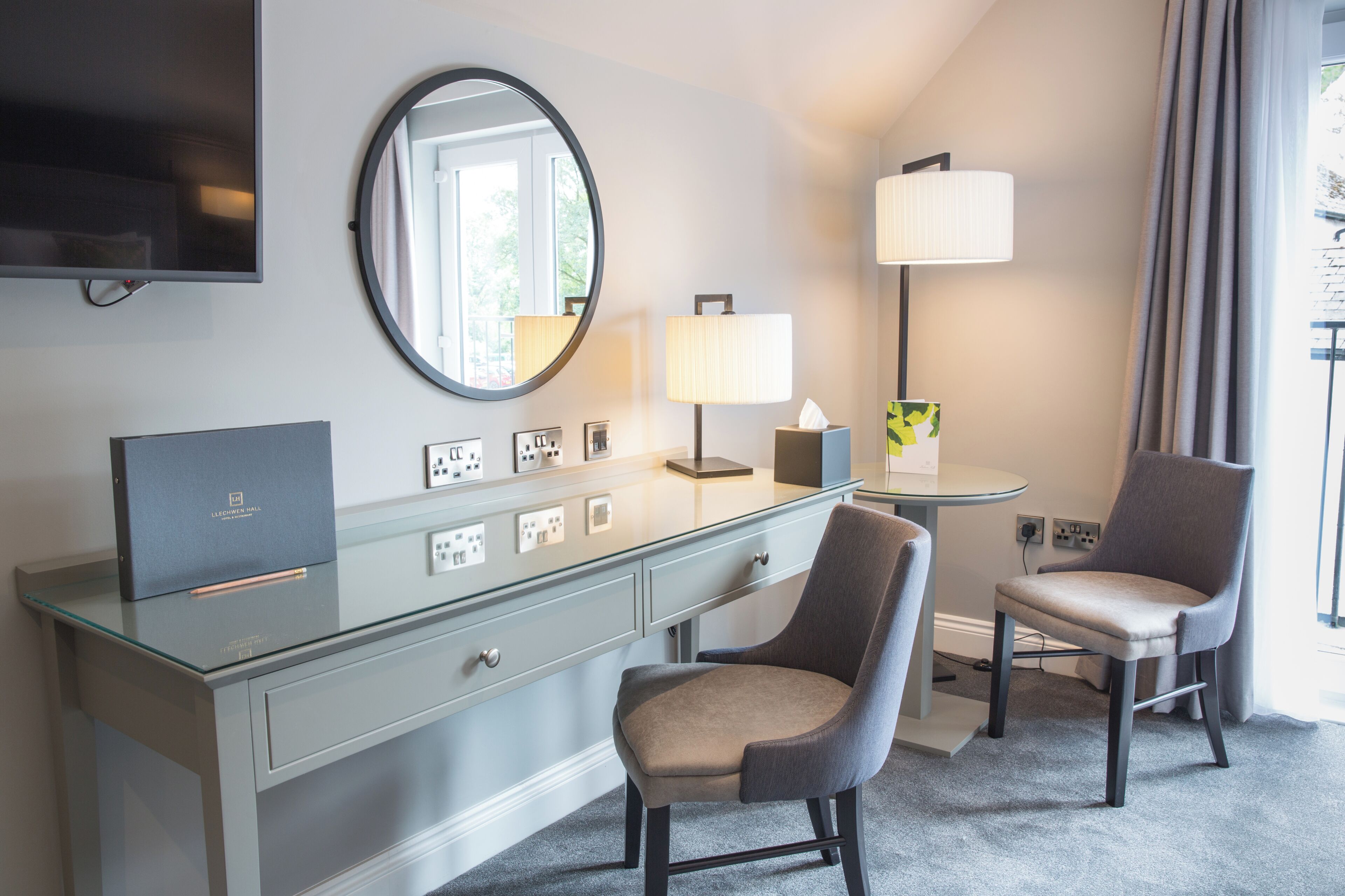 A hotel room with a flat-screen TV, a round mirror, a desk with a lamp, two chairs, and a small table.