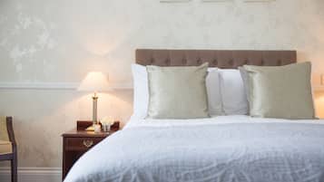 A neatly made bed with a patterned bedspread, a wooden nightstand with a lamp and a vase of flowers, and a chair to the left.