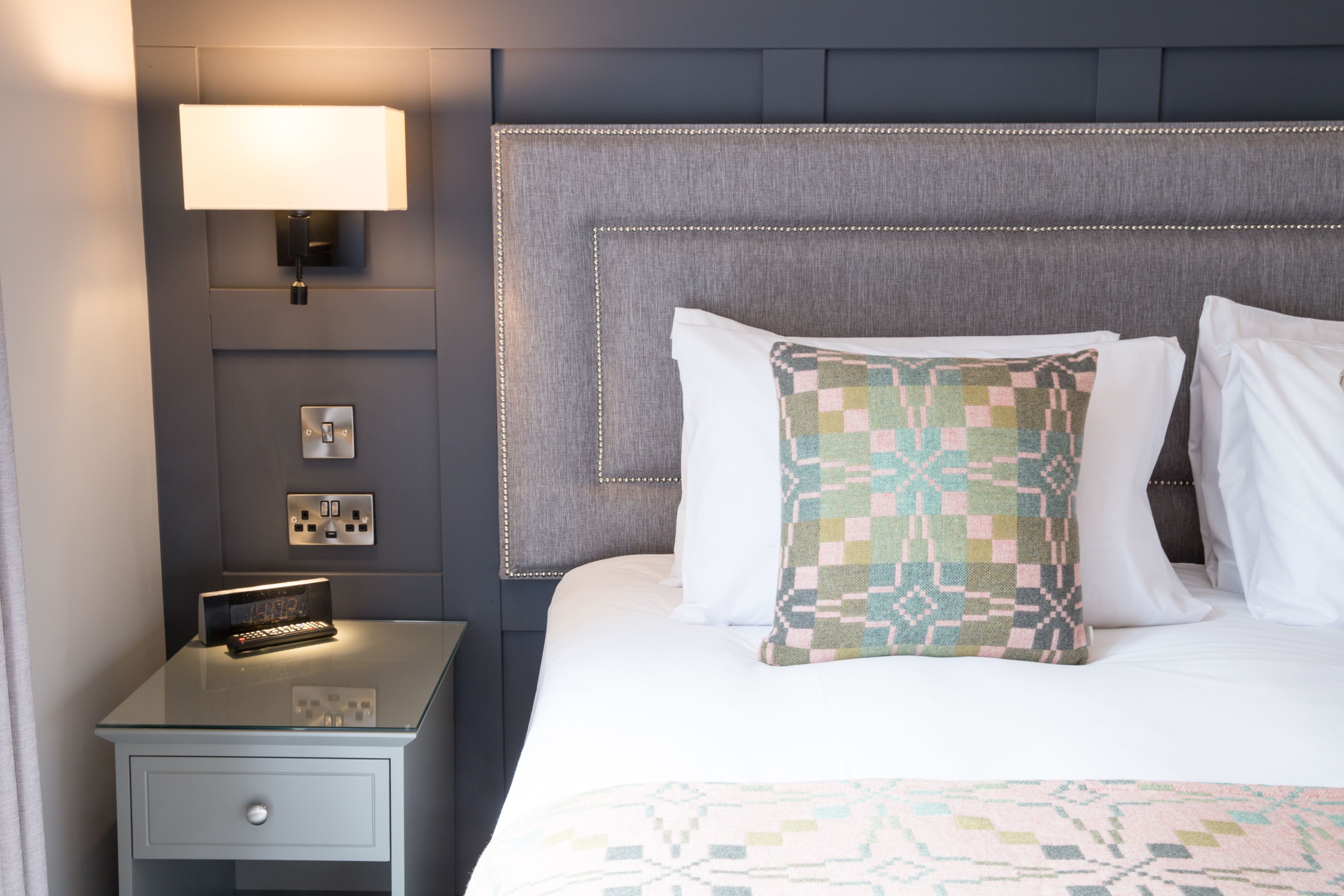 A neatly made bed with a patterned pillow, a bedside lamp, and a nightstand with a small electronic device.