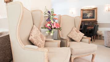 A room with two beige armchairs with floral cushions, a small table with a vase of flowers, and a dining area with a table and chairs in the background.
