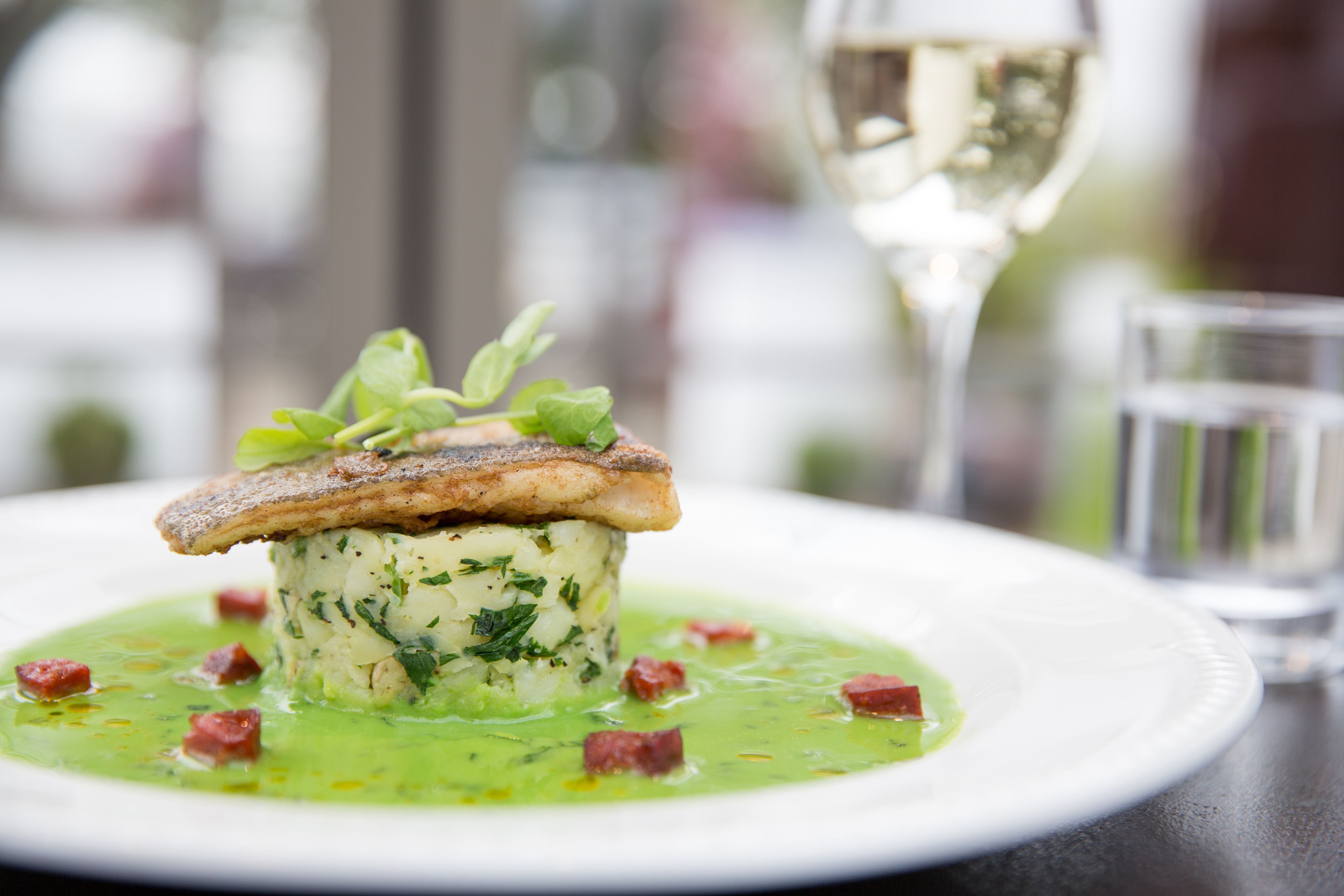 A dish with a fish fillet on top of a creamy base, garnished with herbs, served on a white plate with a green sauce and small red pieces, accompanied by a glass of white wine and a water glass.
