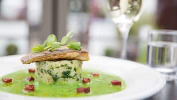 A dish with a fish fillet on top of a creamy base, garnished with herbs, served on a white plate with a green sauce and small red pieces, accompanied by a glass of white wine and a water glass.