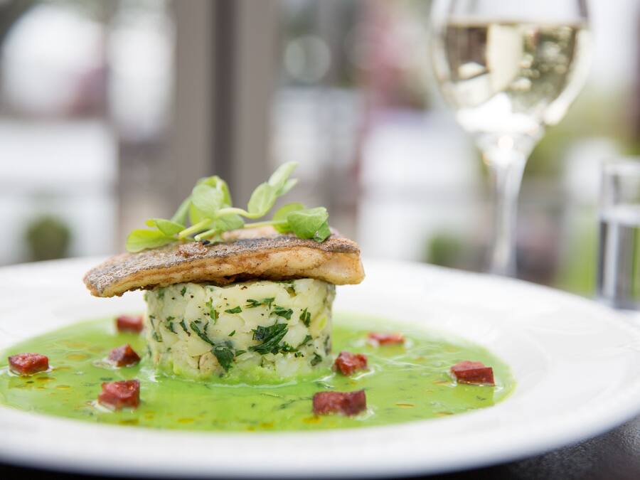 A dish with a fish fillet on top of a creamy base, garnished with herbs, served on a white plate with a green sauce and small red pieces, accompanied by a glass of white wine and a water glass.