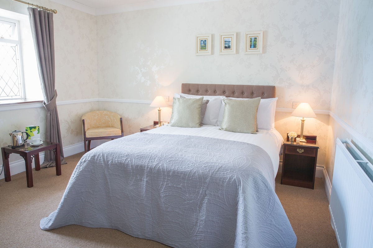 Une chambre à coucher bien rangée, avec un grand lit, deux tables de chevet avec des lampes, une chaise et une petite table sur laquelle se trouvent une bouilloire et une tasse.