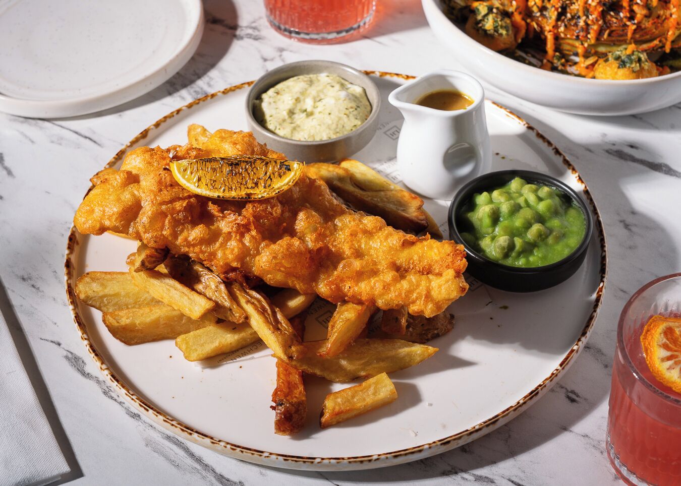 Un plato de pescado con patatas fritas acompañado de guisantes blandos, salsa tártara y un pequeño recipiente con salsa gravy.