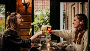 Two people toasting with drinks at a wooden table with a sign reading "LONG LIVE NIN". - Long John’s Hotel (Amersfoort)