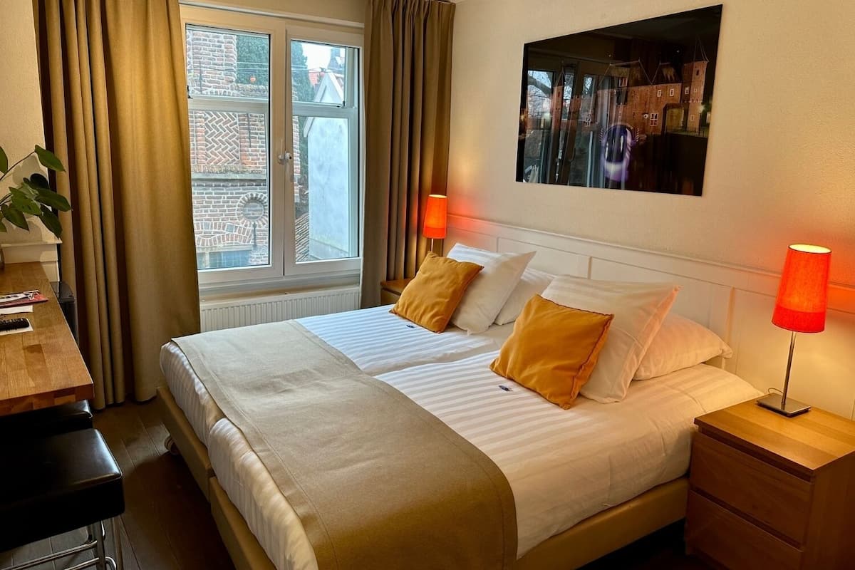 Habitación de hotel con cama, dos lámparas de noche, escritorio con silla, ventana con cortinas y un cuadro en la pared.