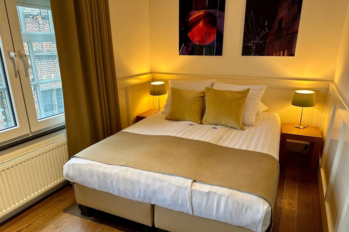 Una cama bien hecha con sábanas blancas y un plaid color beige, con dos mesitas de noche con lámparas, en una habitación con piso de madera y una ventana con cortinas.