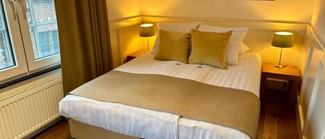 A neatly made bed with white linens and a beige throw blanket, flanked by two bedside tables with lamps, in a room with wooden flooring and a window with curtains.