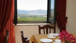 Un coin repas avec une table dressée pour deux personnes, une fenêtre donnant sur un paysage verdoyant et un vase rempli de fleurs roses.
